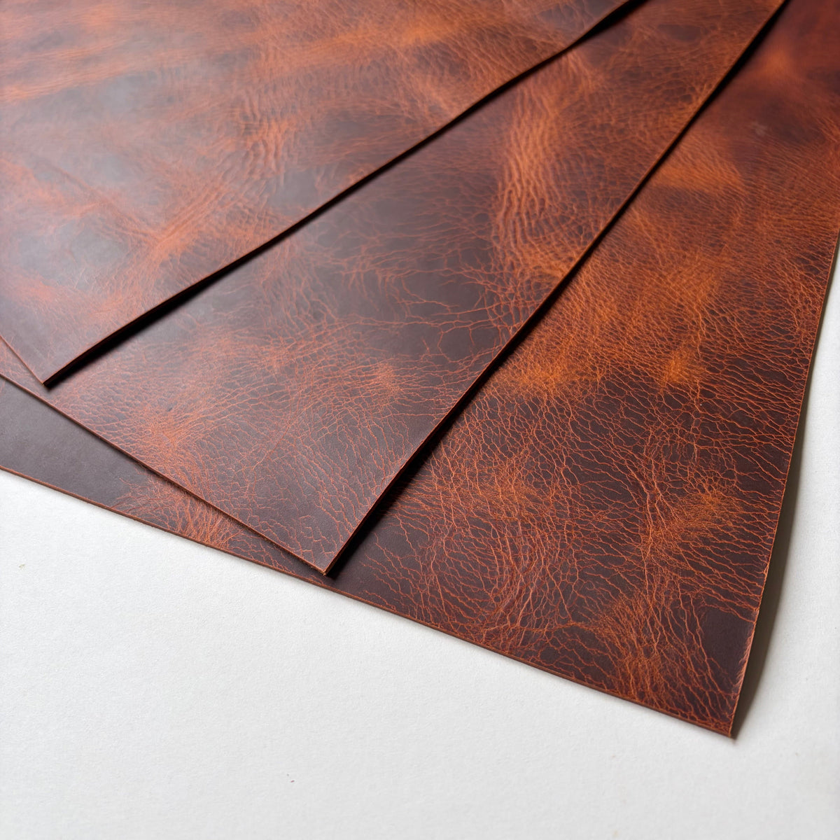 Three textured brown leather sheets with a distressed, cracked grain pattern, arranged diagonally on a light grey surface.