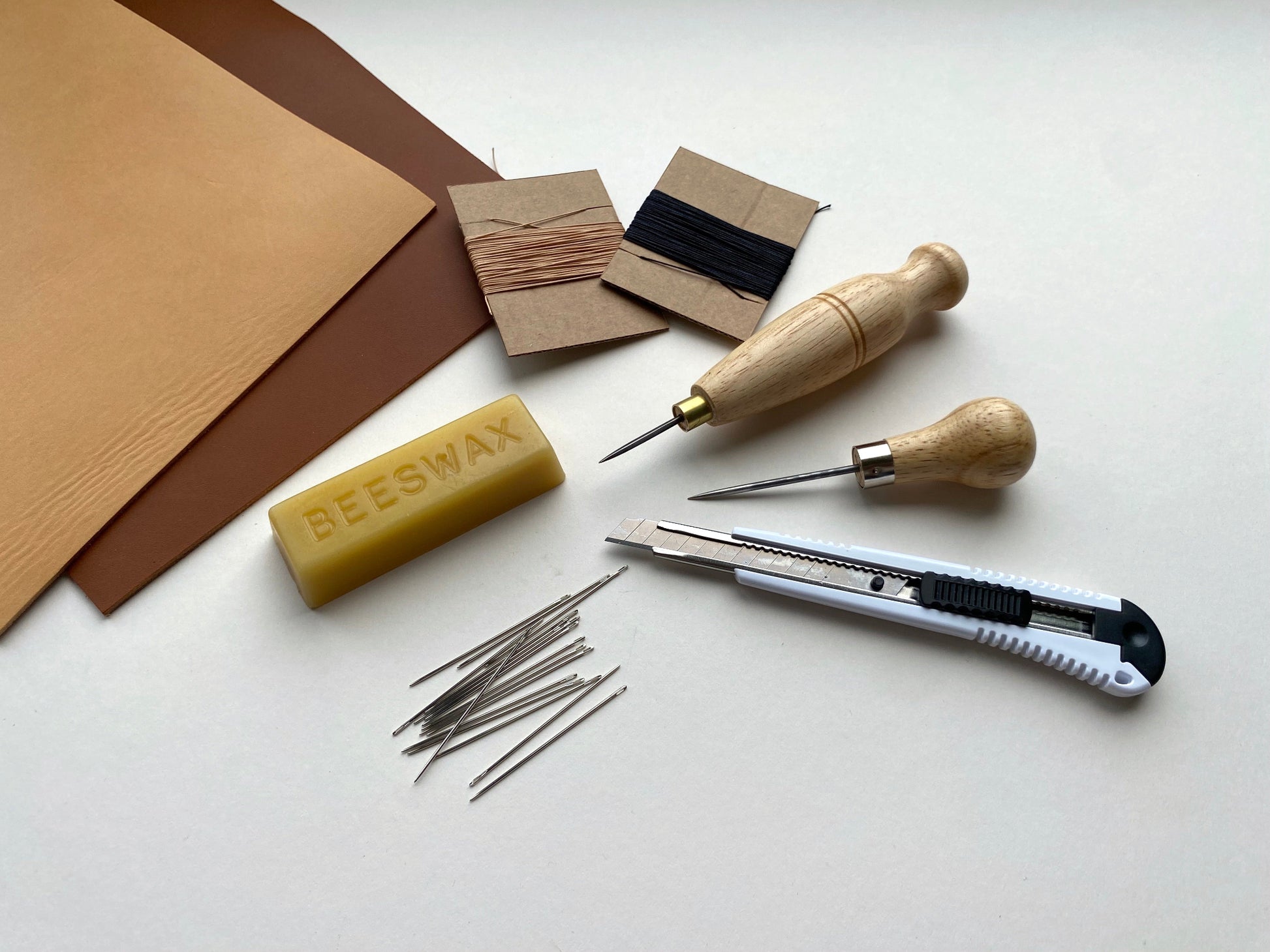 Leatherworking tool kit and materials including beeswax, needles, and a knife on a white surface.
