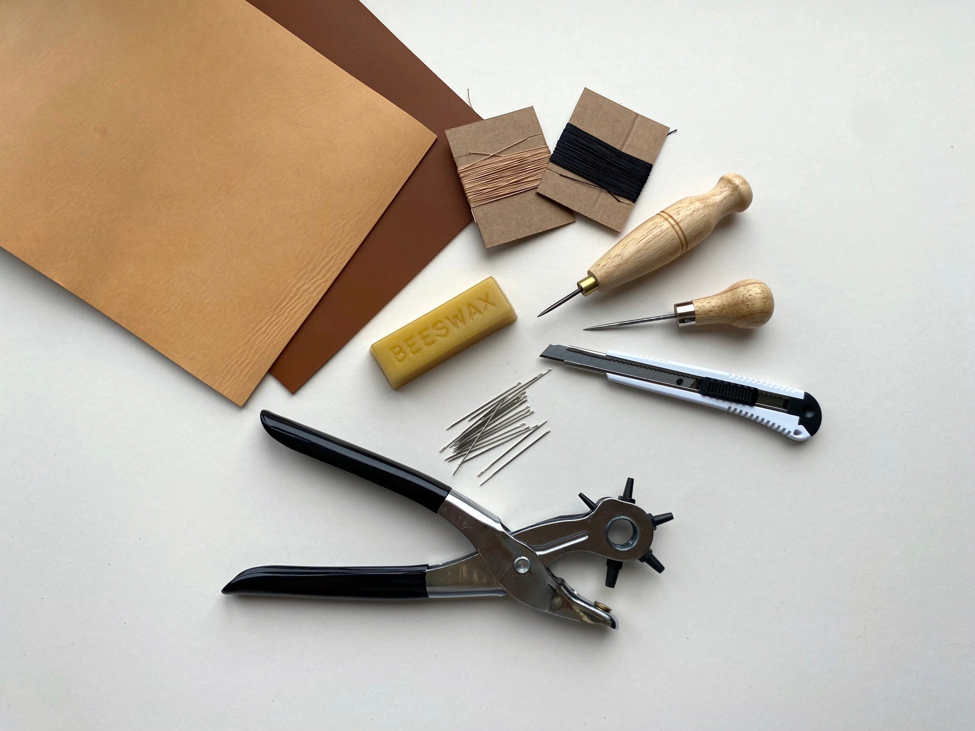 Leatherworking tool kit and materials including beeswax, needles, and a knife on a white surface.