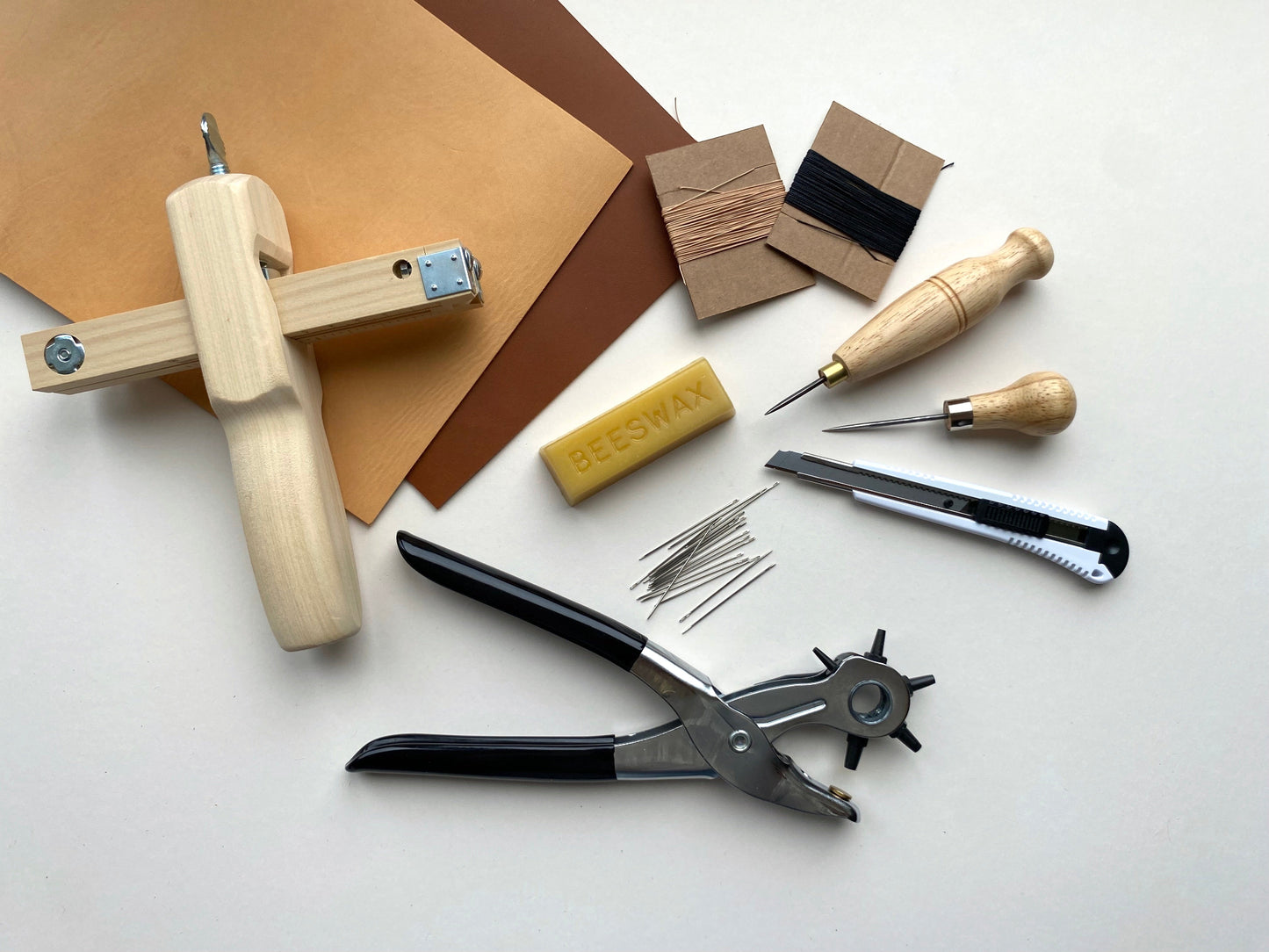 Leatherworking tool kit and materials including beeswax, needles, and a knife on a white surface.