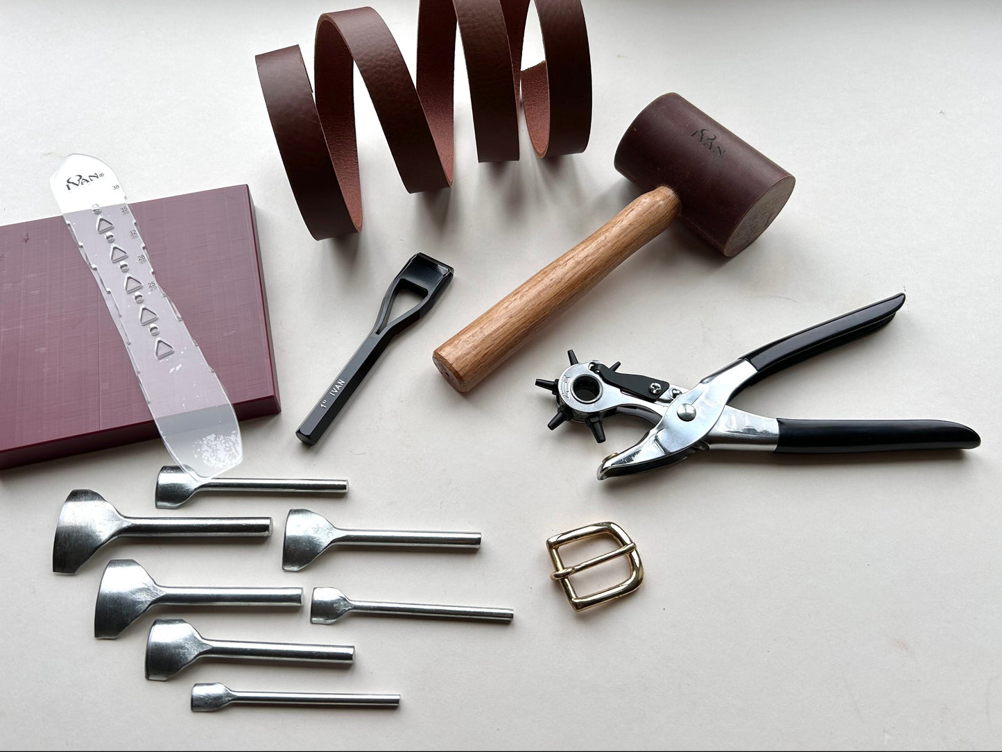 Collection of leatherworking tools including a mallet, springs, pliers, and metal tools on a white surface.