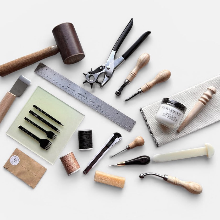 A flat-lay arrangement of leatherworking and crafting tools on a light gray background, including a mallet, ruler, punches, burnishing stones, a small jar, needles, and other hand tools.