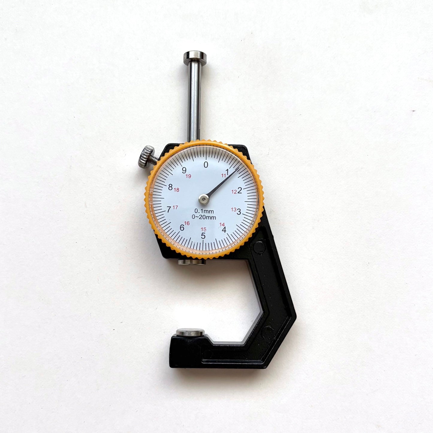 A dial micrometer caliper with a black body, silver metal jaws, and a white face with a yellow-orange bezel, photographed on a plain white background.