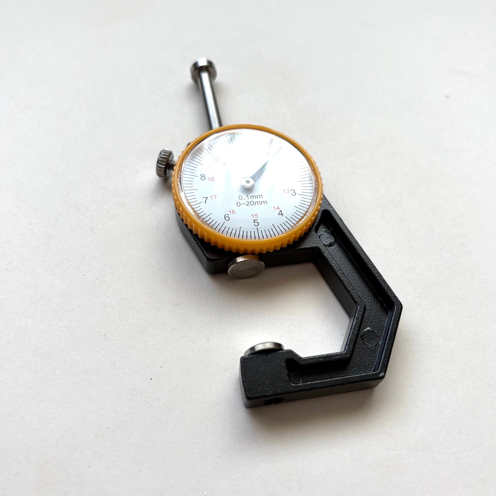 A black precision micrometer caliper with a white dial face and a yellow-orange ring around the scale, photographed on a plain light background.