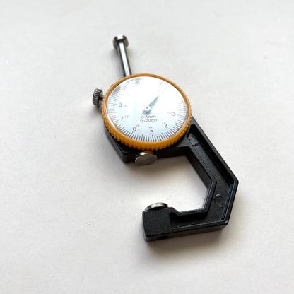 A black precision micrometer caliper with a white dial face and a yellow-orange ring around the scale, photographed on a plain light background.