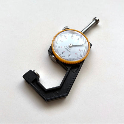 A digital micrometer caliper with a black body, silver metal spindle, and a white dial face with a yellow-orange bezel, shown on a plain off-white background.