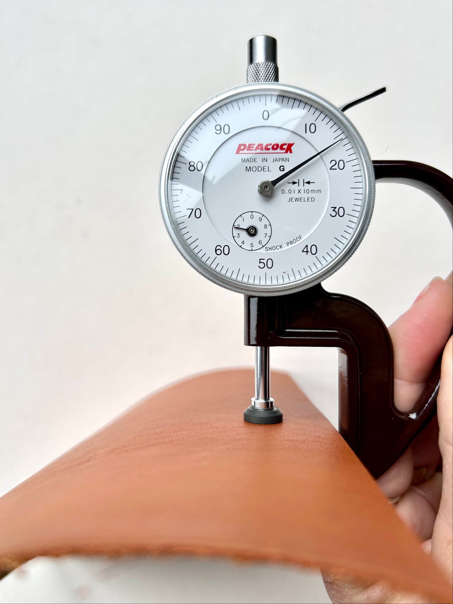 A Peacock dial indicator gauge being used to measure the thickness of a tan leather sheet, held in place by a hand.