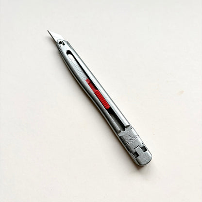 A silver Glanz utility knife with a red sliding scale indicator, shown diagonally on a plain off-white background.