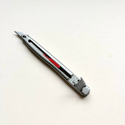 A silver Glanz utility knife with a red sliding scale indicator, shown diagonally on a plain off-white background.
