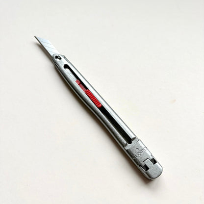 A silver Glanz utility knife with a red sliding scale indicator, shown diagonally on a plain off-white background.