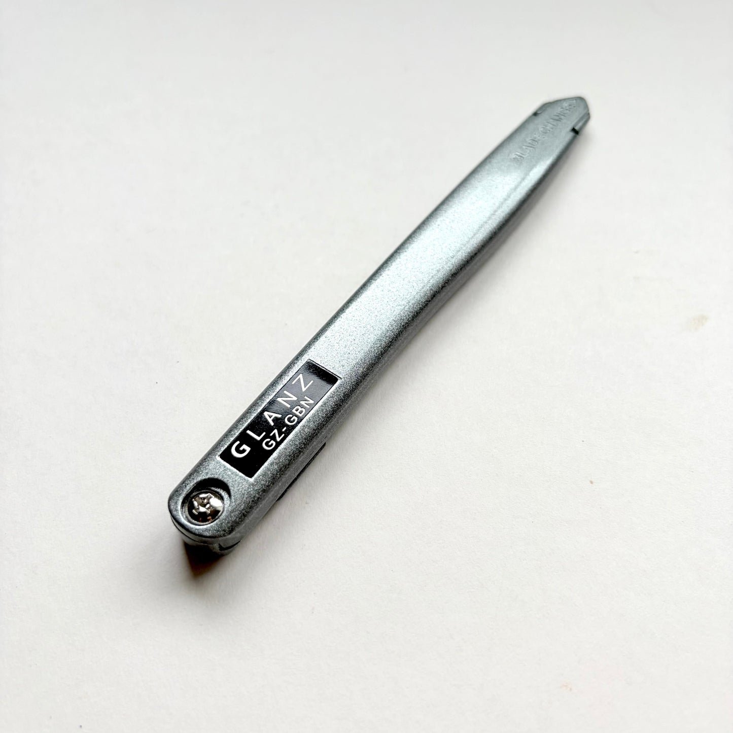A silver metallic pocket knife with a black label that reads 'GLAZ GLOBAL' in white text, photographed diagonally on a plain off-white background.