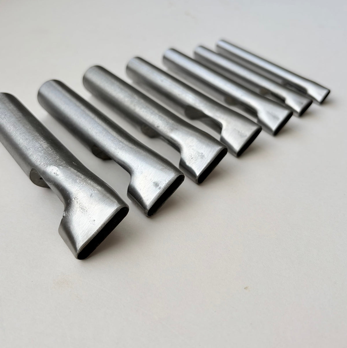 Six metallic silver metal punch tools arranged diagonally on a plain off-white surface.