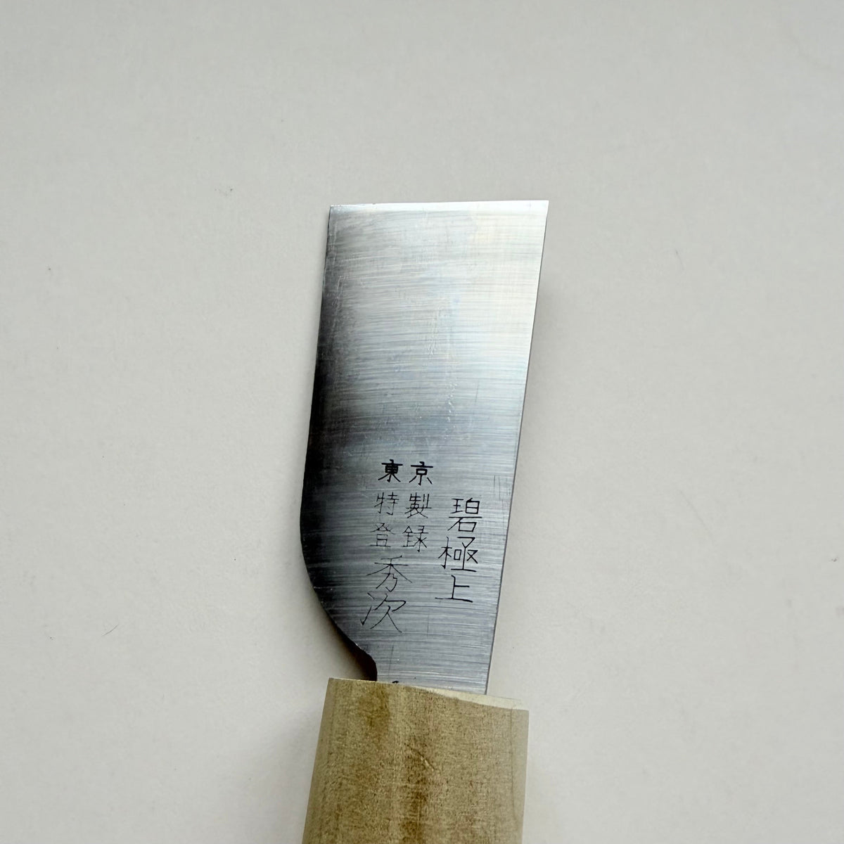 A close-up of a Japanese-style leather paring knife with a satin-finished steel blade and a light-coloured wooden handle, featuring black engraved kanji characters on the blade, set against a plain light grey background.