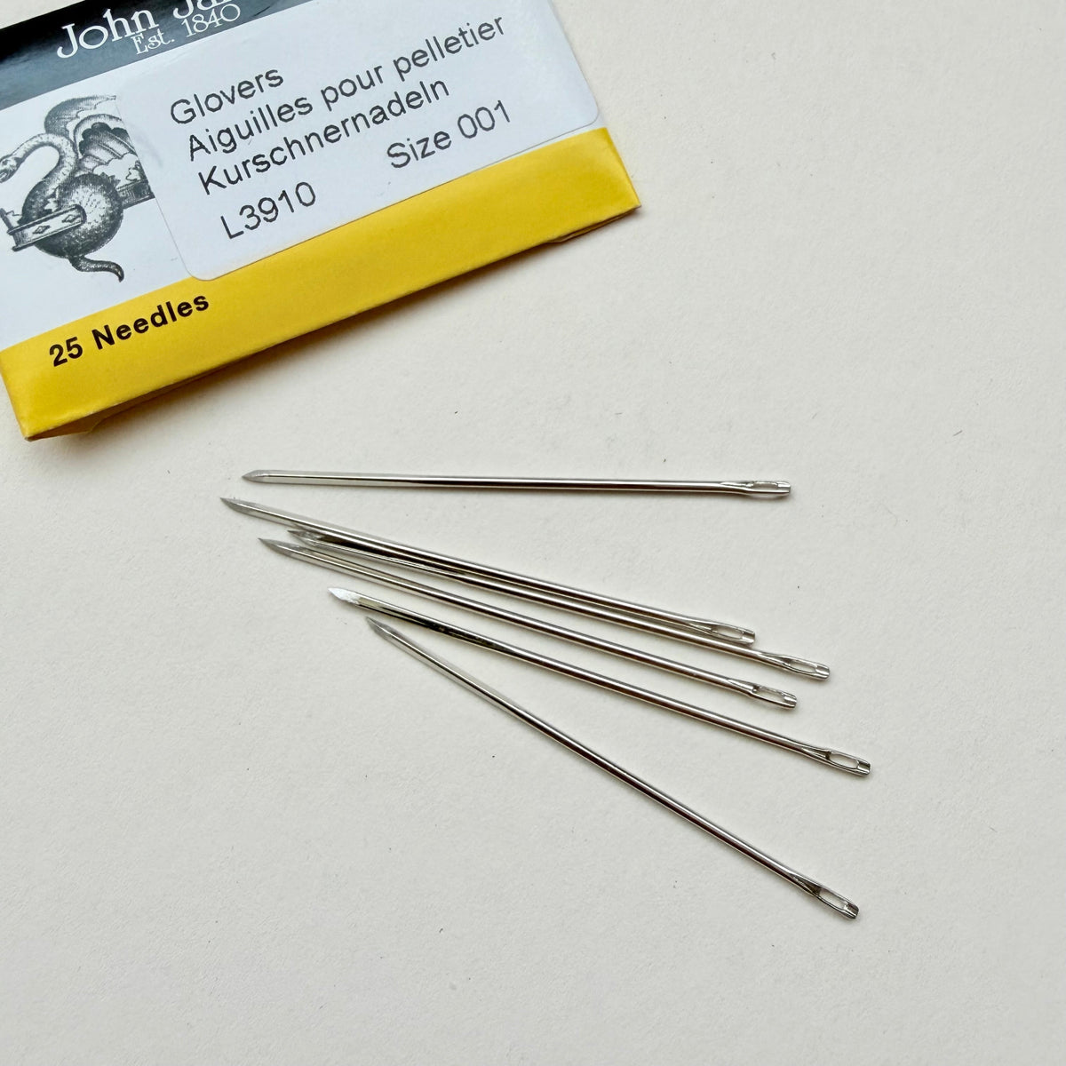 Six metal glove-pellet needles from John James are scattered on a light beige surface, with their yellow and white packaging box visible in the top-left corner.