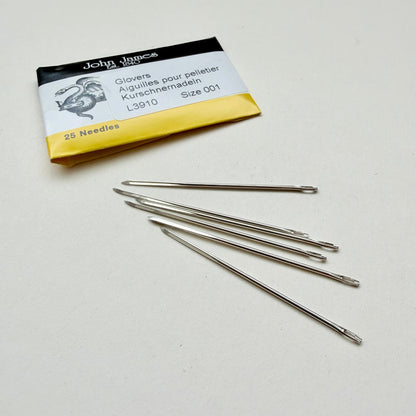 Five metal glove-pellet needles from John James size 001 lie on a light gray surface next to their original packaging card.