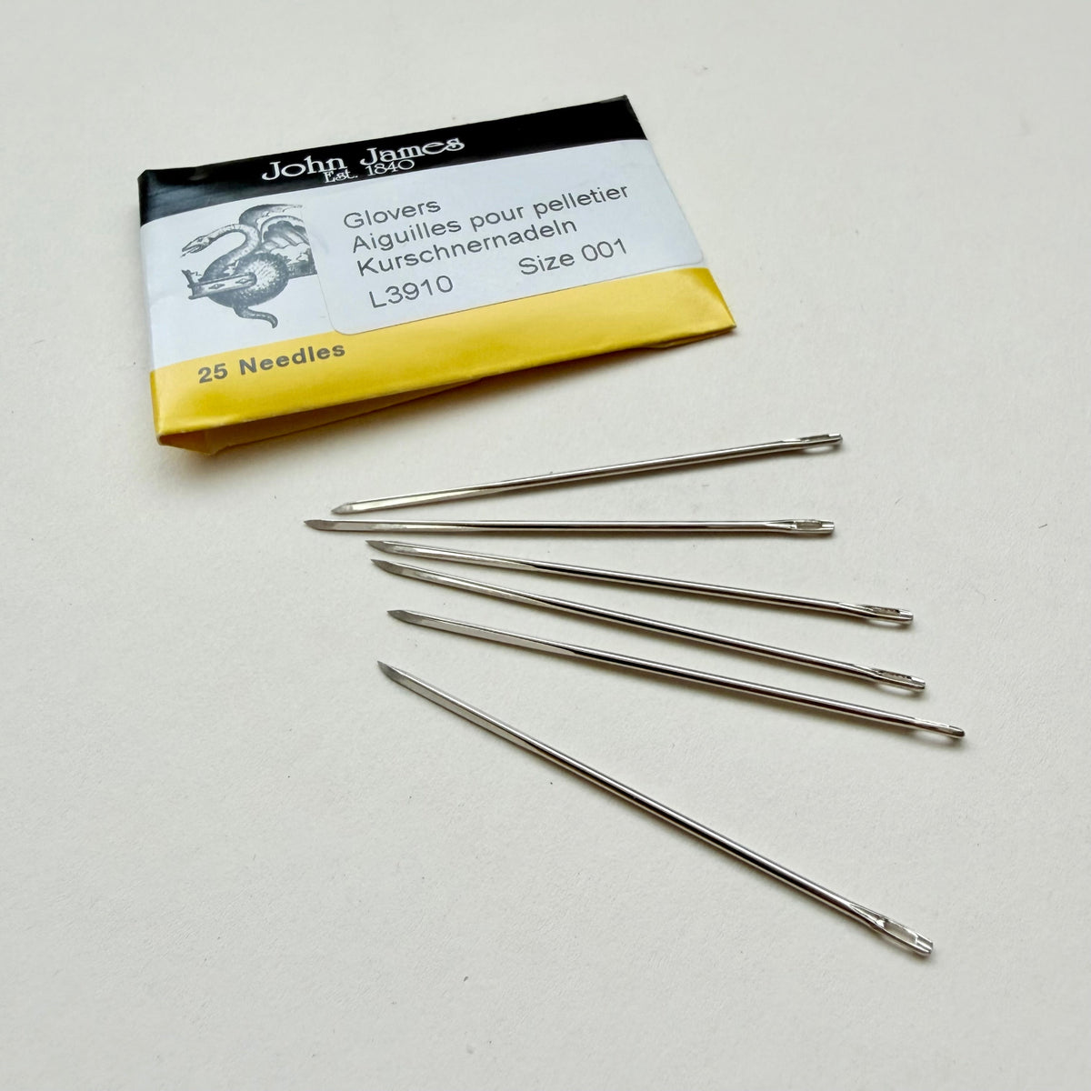 Six silver metal glovers' needles arranged on a light beige surface next to their original John James packaging card.