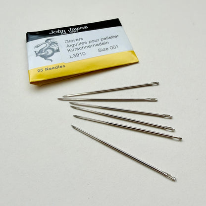 Six silver metal glovers' needles arranged on a light beige surface next to their original John James packaging card.