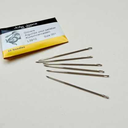 Five metal sewing gloves needles arranged on a light beige surface next to their original yellow and black John Jones packaging.