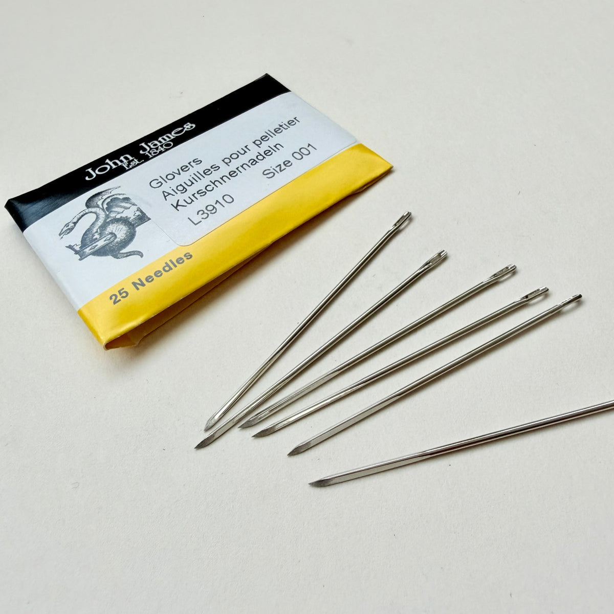 Five metal glover needles arranged on a light surface beside their branded paper packaging, which reads John James and shows size 001, 25 needles.