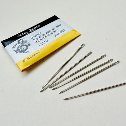 Five metal glover needles arranged on a light surface beside their branded paper packaging, which reads John James and shows size 001, 25 needles.
