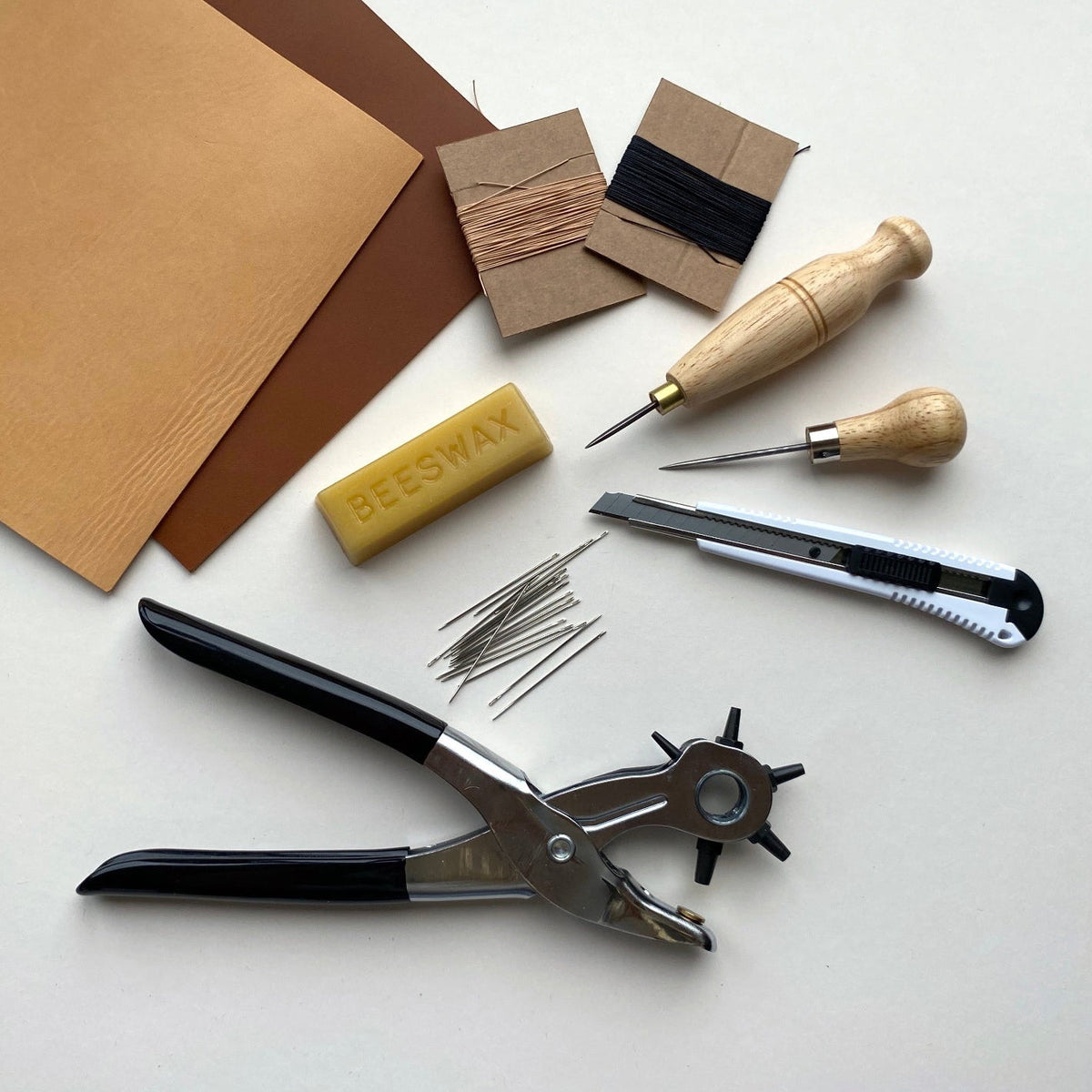 Leatherworking tool kit and materials including beeswax, needles, and a knife on a white surface.