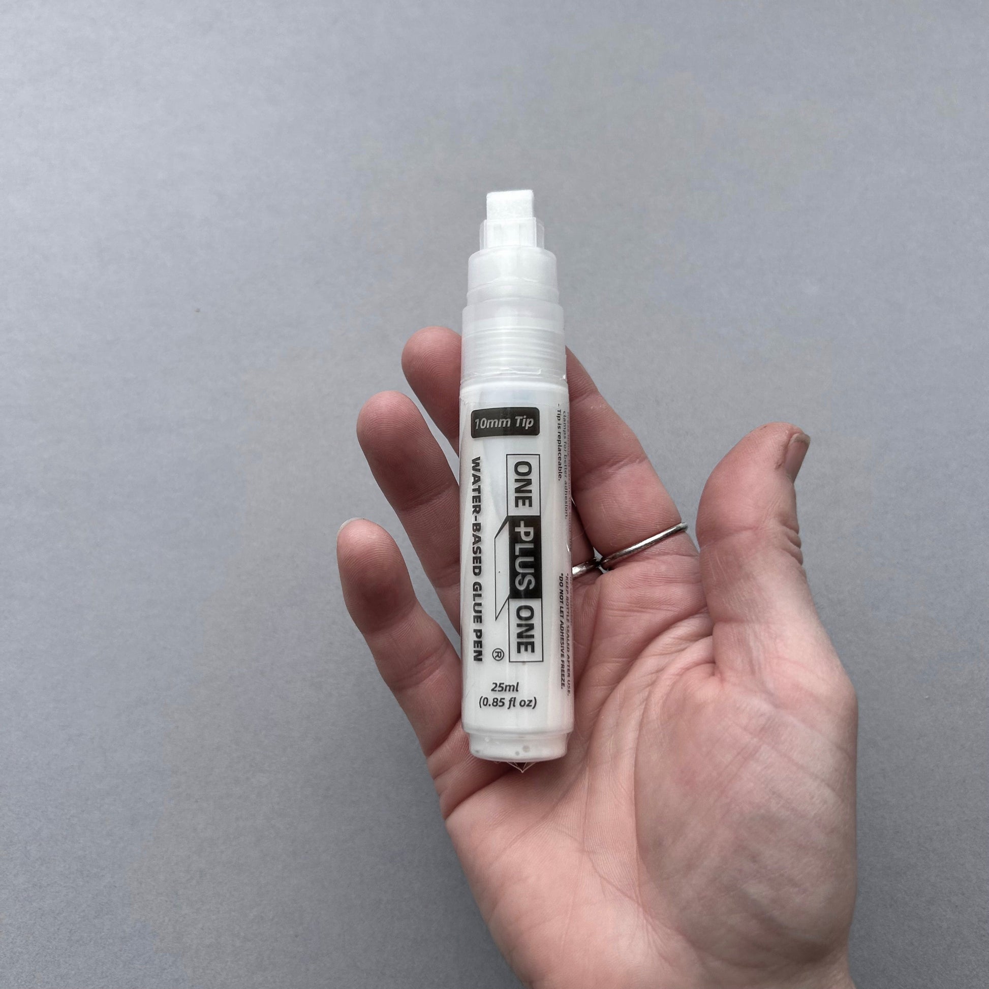 Hand holding a white leather glue pen labeled 'ONE PLUS ONE' against a gray background