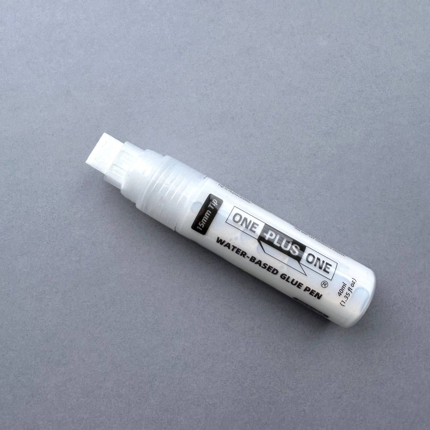 White water-based glue pen labeled 'ONE PLUS ONE' on a gray background