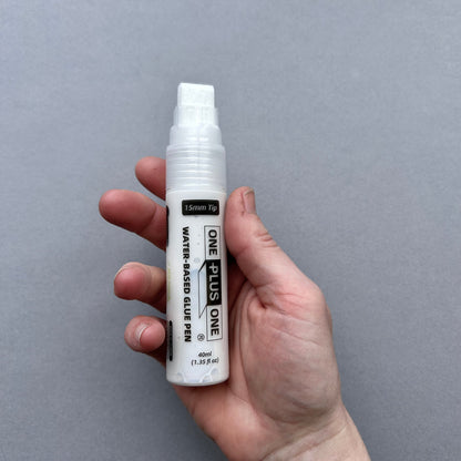 Hand holding a white glue pen labeled 'One Plus One' against a gray background