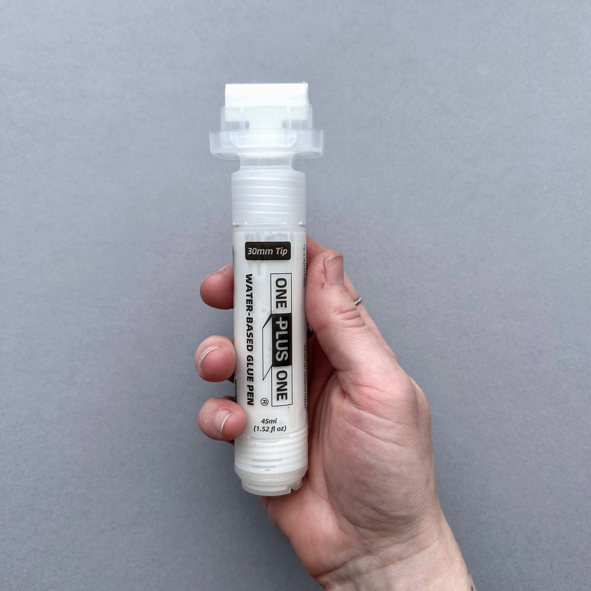 Hand holding a white leather glue pen with 'ONE+ONE' branding against a gray background