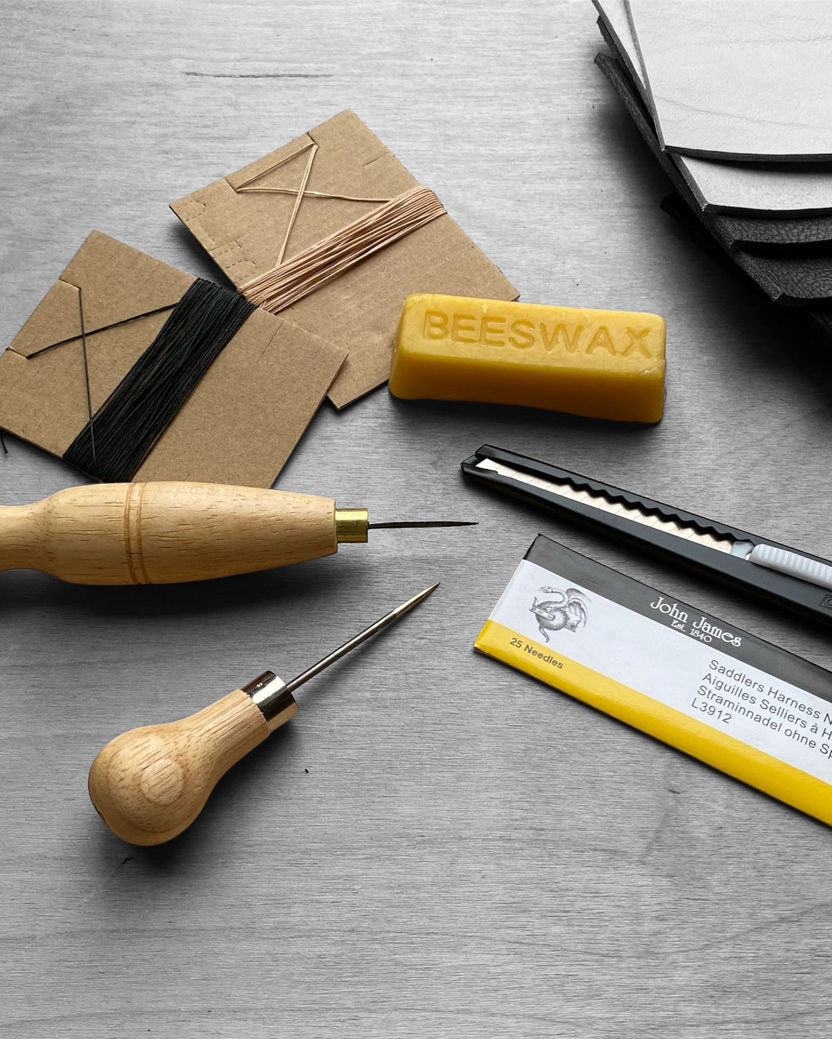 A leatherworking and carving kit arranged on a gray textured surface, including two wooden-handled tools, a yellow block of beeswax, a black utility knife, a card with branding, and several brown leather strips with black cord.