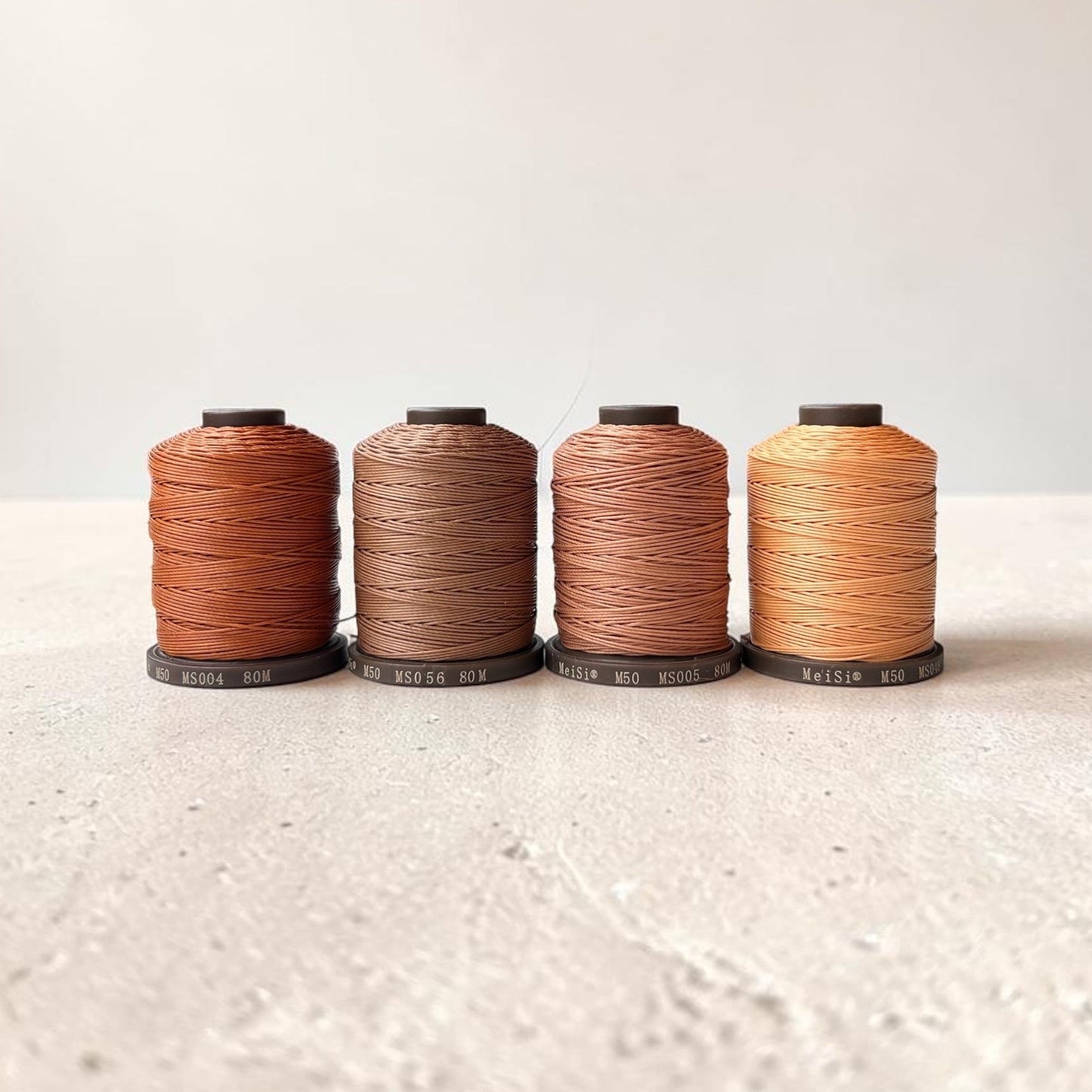 Four spools of Meisi Super Fine Linen thread in different colors on a textured surface with a light background