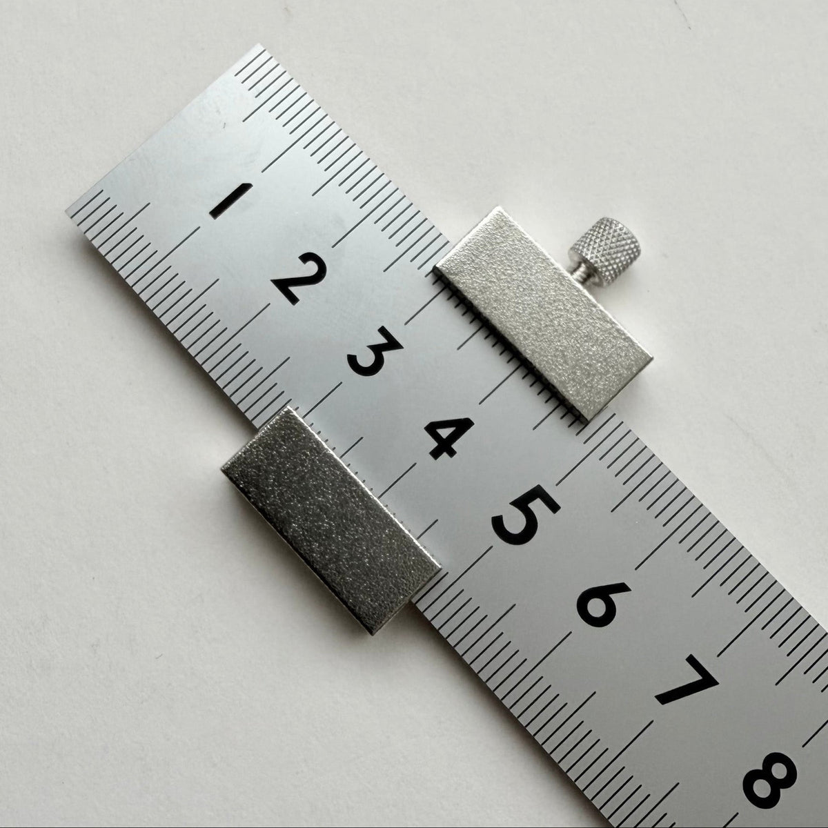 A silver metal ruler with black measurement markings and numbers, shown diagonally on a plain light gray background.