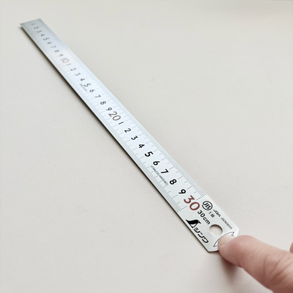 A stainless steel ruler with Japanese markings and a hanging loop, held by a hand on a plain off-white surface.