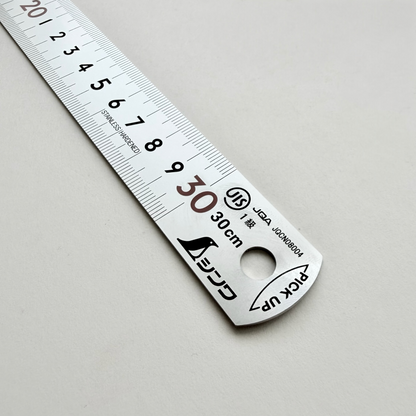 A stainless steel ruler with black printed measurements and Japanese text, photographed diagonally on a light gray background.