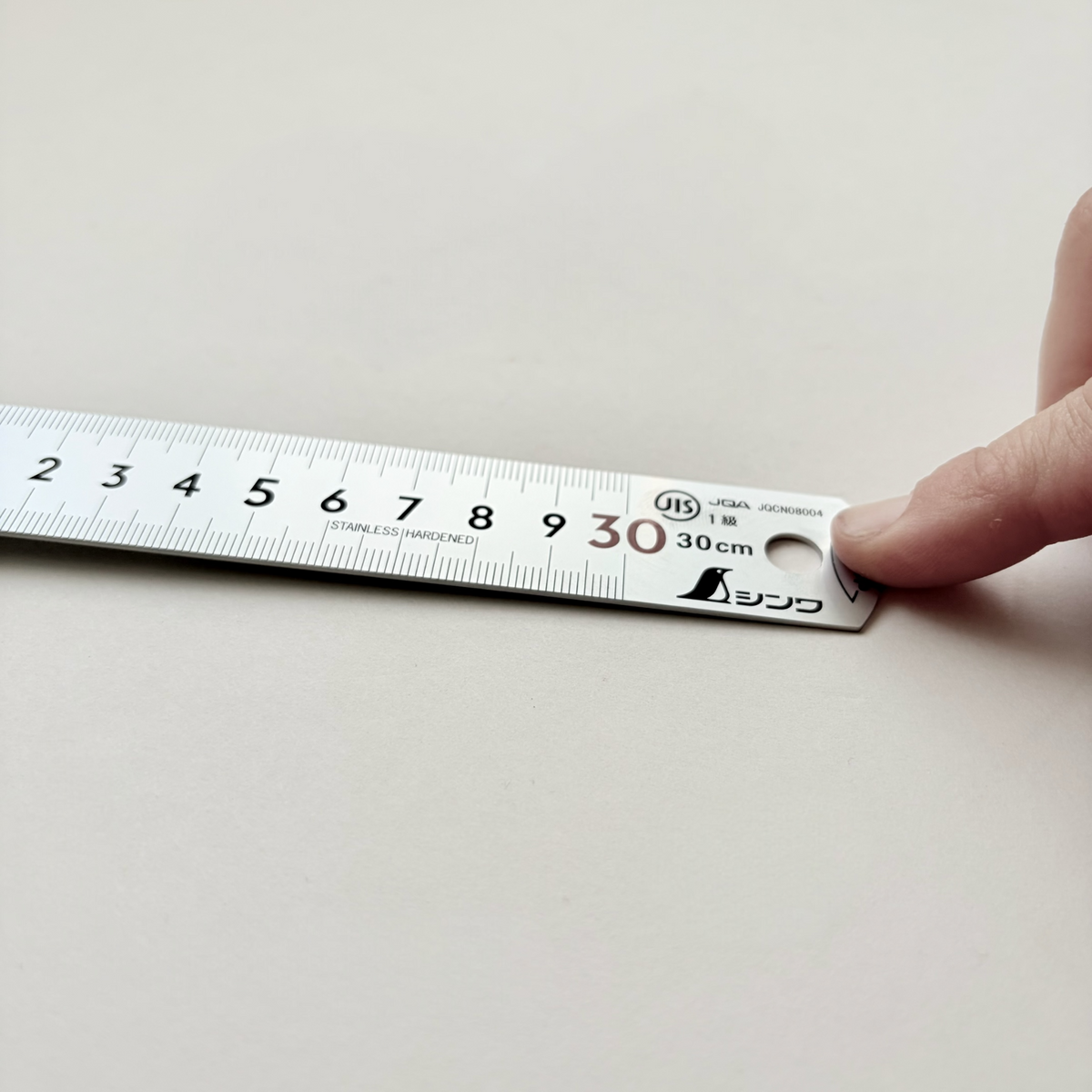 A 30 cm stainless steel ruler with Japanese markings and a keyhole hole, held by a finger on a light beige surface.