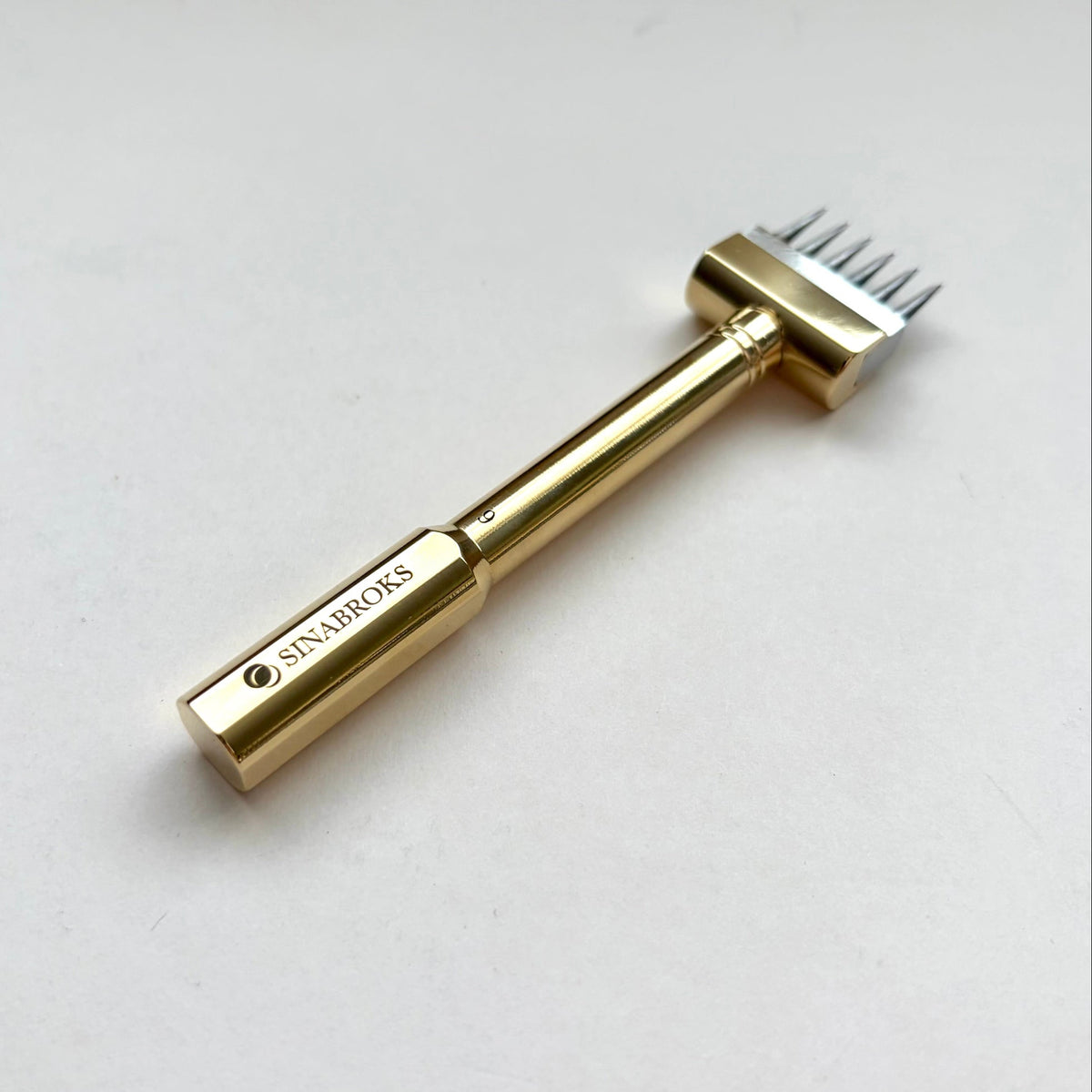 A gold-colored metal hair removal tool with a multi-pronged head, featuring the brand name 'SINABROKS' engraved on the handle, set against a plain light gray background.