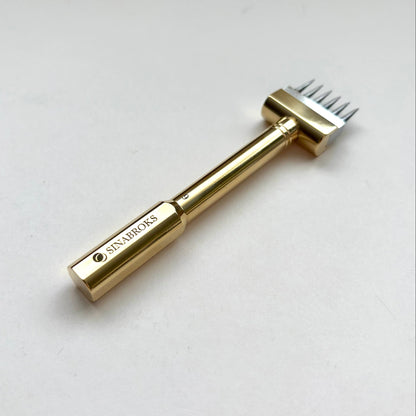 A gold-colored metal hair removal tool with a multi-pronged head, featuring the brand name 'SINABROKS' engraved on the handle, set against a plain light gray background.