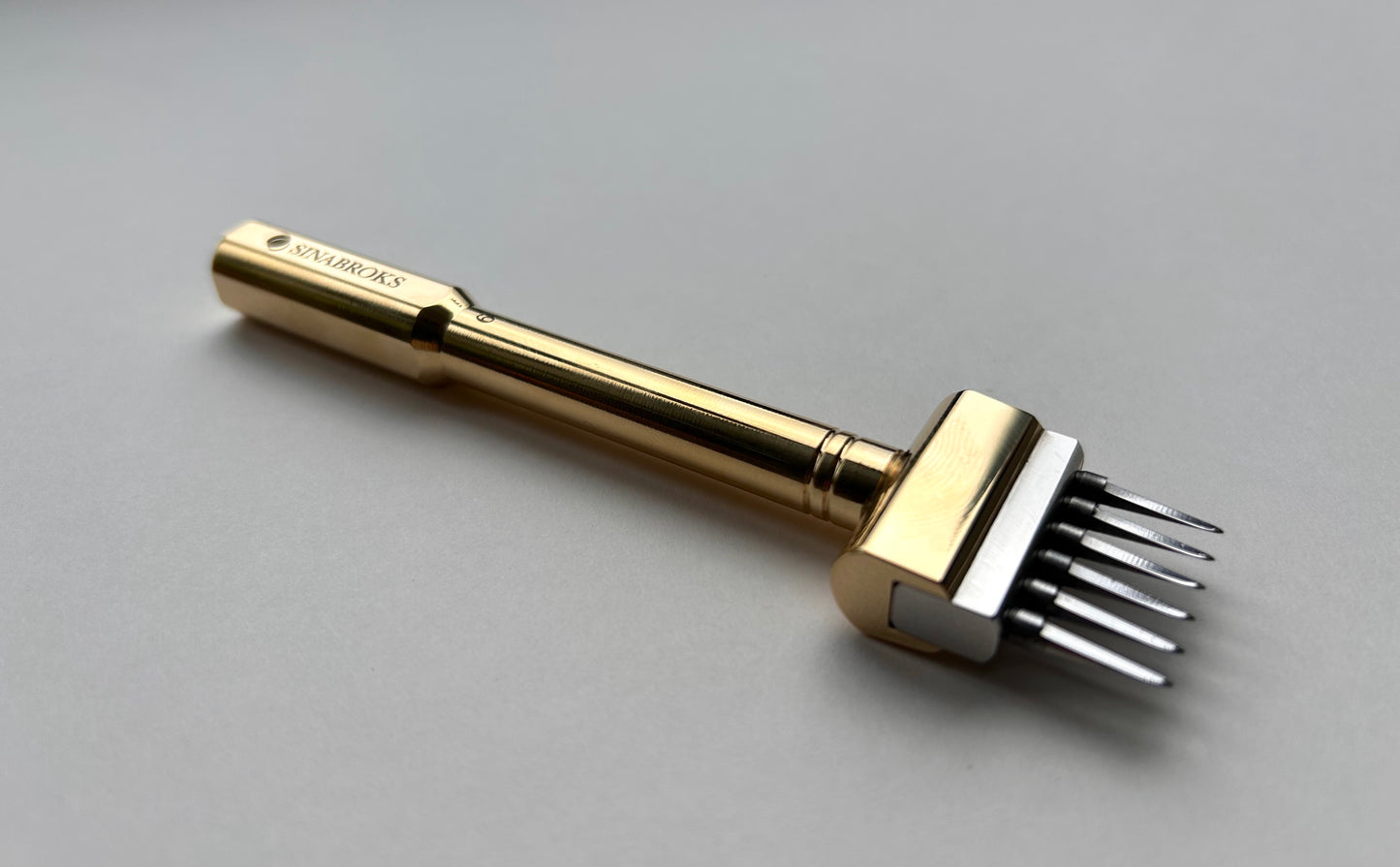 Gold-colored Sinabroks leather pricking iron tool on a light gray background side profile