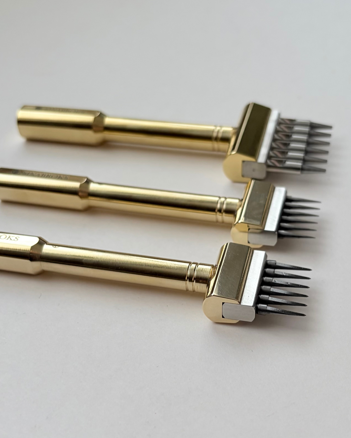 Three gold-coloured metal sinabroks leather punches arranged diagonally on a light grey background.